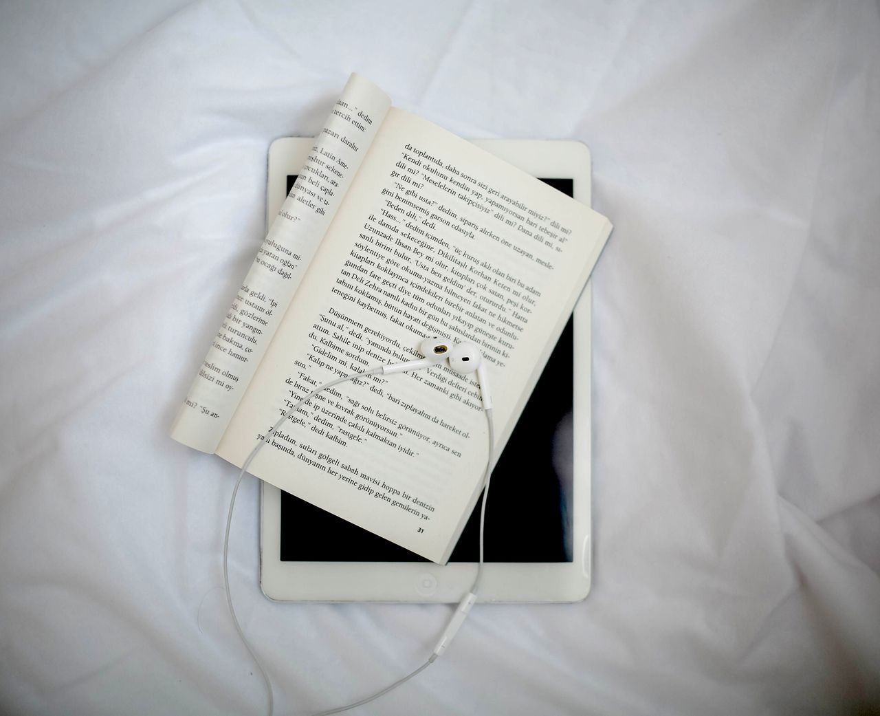 Open book with earphones on a tablet placed on white fabric, from above.
